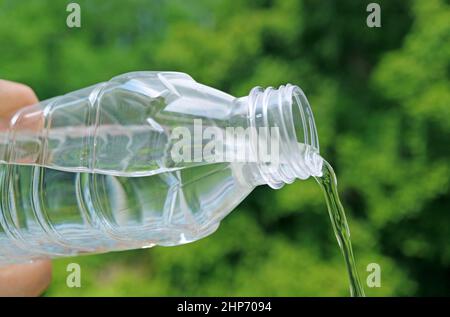 Trinkwasser wird aus einer Plastikflasche mit verschwommenem grünem Laub im Hintergrund gegossen Stockfoto