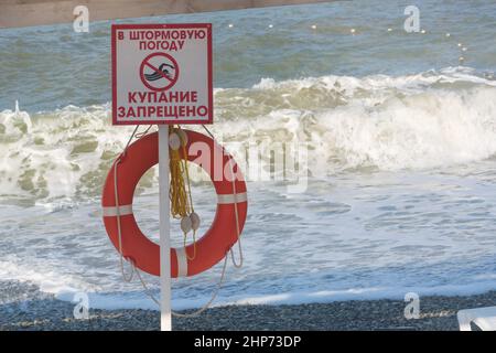 Schild mit der Aufschrift auf Russisch „bei stürmischem Wetter ist es verboten zu schwimmen“ vor dem Hintergrund großer Wellen und Rettungsboje. Verbotene Zone auf dem Stockfoto