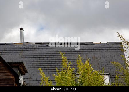 Ein Hausdach mit beschädigten und fehlenden Bergkacheln, das an einem stürmischen Tag mit bewölktem Himmel abgebildet ist. Eine Bambushecke bläst im Vordergrund. Stockfoto