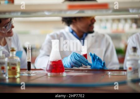 Junge Kollegen in Schutzkleidung diskutieren während der Arbeit mit bunten Chemikalien in entspannter Atmosphäre im Universitätslabor. Wissenschaft, Chemie Stockfoto