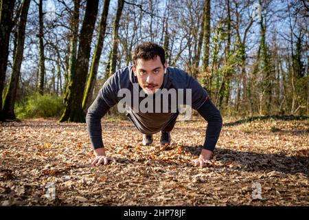 Der Läufer macht Liegestütze im Wald. Der junge Mann mit den blauen Augen und dem Schnurrbart macht Liegestütze auf dem Boden, während er der Musik zuhört Stockfoto