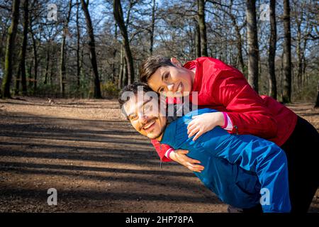 Das junge Paar spielt während eines Trainings glücklich im Wald. Der gutaussehende Mann trägt seine Freundin auf dem Rücken. Stockfoto