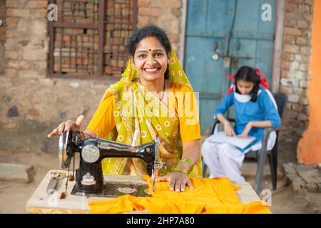 Porträt einer glücklichen, traditionellen indischen Frau, die mit einer Nähmaschine einen Sari trägt, während ihre junge Tochter hinter ihr in Schuluniform studiert. Stockfoto