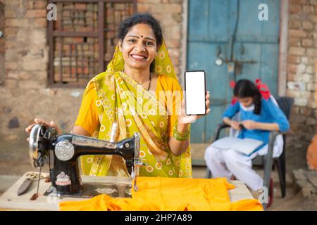 Porträt einer glücklichen traditionellen indischen Frau, die mit einer Nähmaschine einen Sari trägt, während sie ein Smartphone mit leerem Display zeigt, um eine leere Anzeige zu platzieren Stockfoto