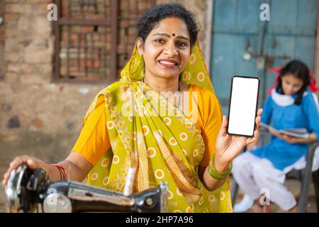 Porträt einer glücklichen traditionellen indischen Frau, die mit einer Nähmaschine einen Sari trägt, während sie ein Smartphone mit leerem Display zeigt, um Werbung zu schalten, weiblich Stockfoto