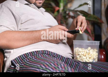 Junger Mann, der Popcorn isst und Popcorn hält, sitzt auf dem Sofa Stockfoto