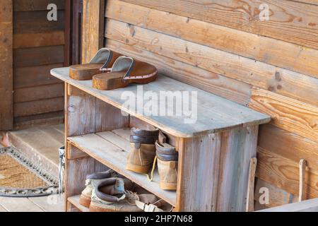 Alte Holzschuhbox in der Nähe eines Holzhauses auf einem Bauernhof. Die Schuhbox hat alte Clogs, Gummistiefel und Schuhe. Stockfoto