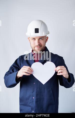Labor Day: Netter männlicher Techniker, Elektriker, Auftragnehmer, Supervisor oder Ingenieur in Hardhat und blauer Uniform mit weißem Papier in Herzform. Konzept des Internationalen Tages der Arbeit. Hochwertige Bilder Stockfoto