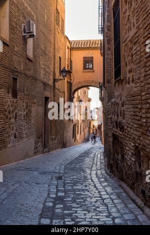 Cobblestone Street - Ein sonniger Abend Blick auf eine Backsteinbrücke über einer alten engen Kopfsteinpflasterstraße im jüdischen Viertel der historischen Stadt Toledo, Spanien. Stockfoto