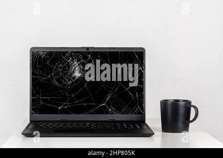 Ein Laptop mit einem kaputten Bildschirm und einem schwarzen Becher Tee auf einem Tisch an einer weißen Wand Stockfoto