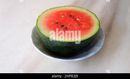 Ganze Wassermelone in zwei Hälften geschnitten und auf einem kleinen Teller serviert. Auf einer Marmoroberfläche. Stockfoto