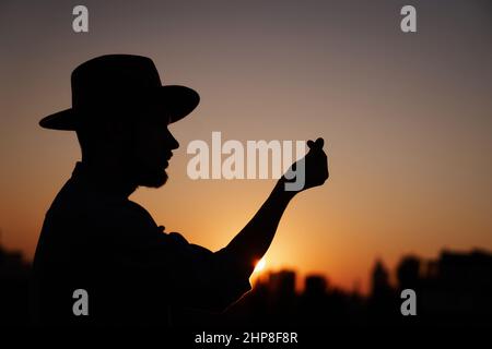 Silhouette eines Mannes im Hut bei Sonnenuntergang, der ein koreanisches Herzzeichen mit Händen zeigt. Hipster-Mann mit „Mini-Herz“-Geste mit Blick auf die Stadt. Liebe oder Romantik Konzept. Hochwertige Bilder Stockfoto