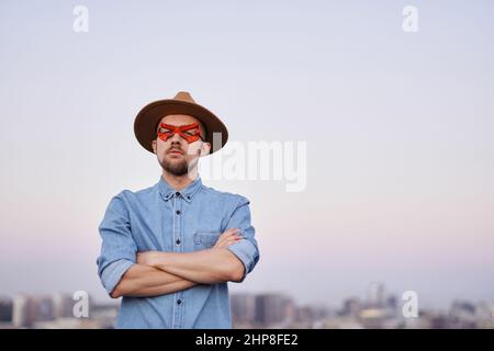 Netter kaukasischer bärtiger Mann als Superheld mit roter Gesichtsmaske und Hut mit gekreuzten Armen. Stark selbstbewusster Mann gegen den Blick auf die Stadt bei Sonnenuntergang. Konzept von Macht, Gerechtigkeit und Stärke Stockfoto