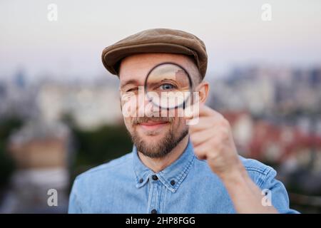 Niedlicher Mann, der bei Sonnenuntergang nach Verkäufen oder Rabatten sucht, mit einer Lupe mit Blick auf die Stadt. Attraktiver bärtiger Mann, der mit einer Lupe auf die Kamera blickt und Jeans-Hemd und Spitzenmütze trägt. Hochwertige Bilder Stockfoto