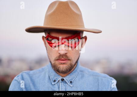 Nahaufnahme Porträt eines bärtigen ernsthaften Mannes in blauer Superheldenmaske, braunem Hut und Jeans-Hemd, das vor der Kamera mit Blick auf die Stadt posiert. Mann Macht oder Gerechtigkeit Konzept. Hochwertige Bilder Stockfoto