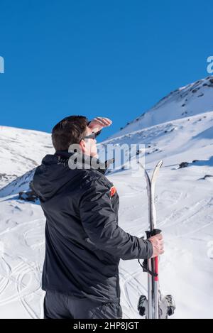 Skifahrer auf dem verschneiten Berg, der seine Augen mit der Hand bedeckt, um an einem sonnigen Tag den Horizont zu sehen Stockfoto