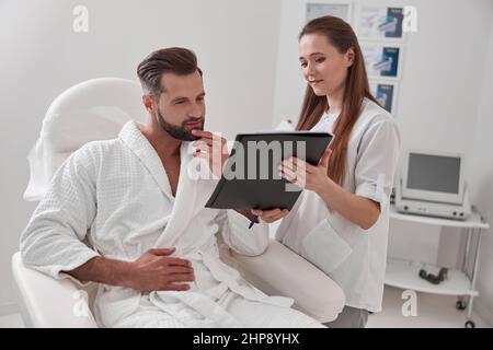 Administrator zeigt Clipboard mit Formular an den Klienten, der in einem Sessel in der kosmetologischen Klinik sitzt Stockfoto