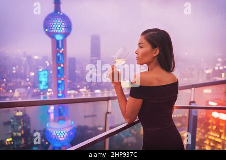 Luxus Shanghai Lifestyle Asiatische Frau trinkt Weißwein auf China Travel. Elegantes Modell in schwarzem Kleid auf der Terrasse mit Blick auf die Stadt, High-End Stockfoto