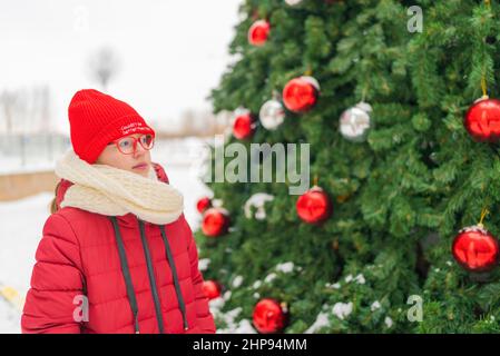 Mädchen in roter Jacke und Hut in der Nähe eines großen Weihnachtsbaums Stockfoto