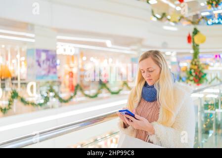 Ein Teenager-Mädchen in einer weißen Jacke in einem Einkaufszentrum, spricht am Telefon vor dem Hintergrund eines Schaufensters mit Rabatten während eines Verkaufs. Stockfoto