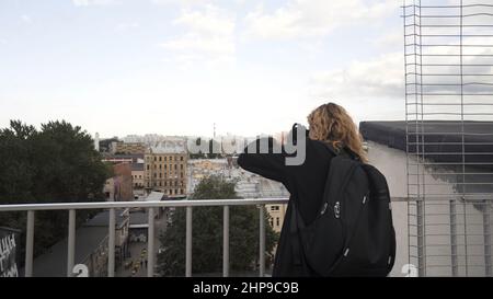 Ziemlich junger Tourist, der während der Stadtbesichtigung Fotos fotografiert. Frau, die auf der Straße spazieren geht und vom Dach aus ein wunderschönes Panorama einer Stadt fotografiert Stockfoto