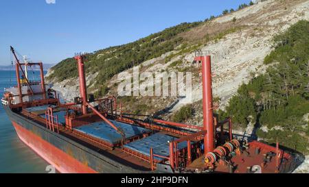 Luftaufnahme eines roten Industrieschiffes mit Menschen an Bord, die Seemannsarbeiten in der Nähe der Küste vertäut haben. Große Barge an der Küste in der Nähe von bewaldeten Hang und pe Stockfoto