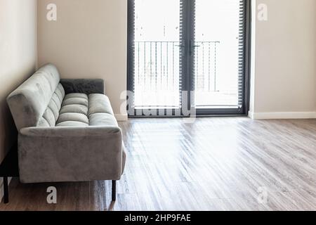 Leeres Wohnzimmer mit Futon Couch graues Sofa im neuen modernen Luxus-Apartment-Haus mit Fenstertüren zum Balkon Holzlaminatboden Stockfoto