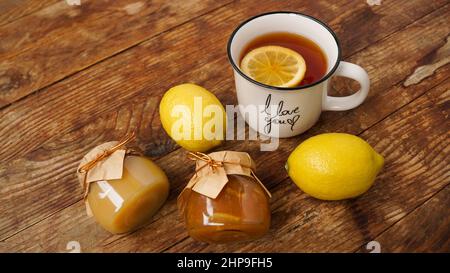 Süßes Frühstück. Tasse schwarzen Tee mit Zitrone. Zitronen und Gläser mit hausgemachter Marmelade, Marmelade oder Quark auf einem Holztisch. Stockfoto
