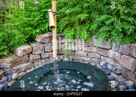 Reinigungsbrunnen in Japan mit Wasser läuft aus Ausguss Wasserhahn im traditionellen japanischen Stil mit grünen Pflanzen und Felswand Garten in Temple Shr Stockfoto