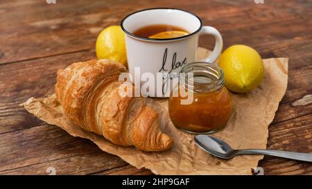 Zitronentee in einer weißen Tasse Ich liebe dich, hausgemachte Marmelade, ganze Zitronen, Croissant auf Holztisch. Zutaten für die Zubereitung von Toast zum leckeren Frühstück Stockfoto
