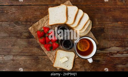 Weiße Tasse schwarzen Tee mit Zitrone. Sandwiches oder Toast mit Butter. Erdbeer- und Marmeladenglas auf Holztisch, Draufsicht. Frühstückskonzept Stockfoto