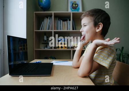 Der 8-jährige Junge sitzt mit einem Laptop am Schreibtisch, ragt seine Zunge heraus und täuscht sich herum Stockfoto