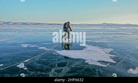 Der Mensch fährt Fahrrad auf dem Eis. Eis des gefrorenen Baikalsees. Rider ist in schwarzer Daunenjacke, Radrucksack, Helm gekleidet. Reifen auf verdecktem Witz Stockfoto