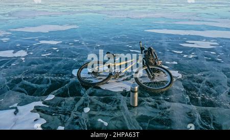 Mann und Fahrrad auf Eis. Er schaut auf das schöne Eis in den Rissen. Blick auf die erste Person auf dem Fahrrad. In der Nähe befindet sich eine Thermoskanne und ein Fahrradrucksack. Eis des Th Stockfoto