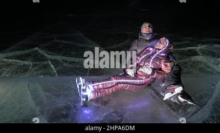 Junge liebende Paar Schlittschuhlaufen auf der Eisbahn in der Nacht. Mann und Frau küssen, umarmen, haben Spaß, lernen zu skaten. Liebe auf Eis machen. Schönes klares Eis mit crac Stockfoto