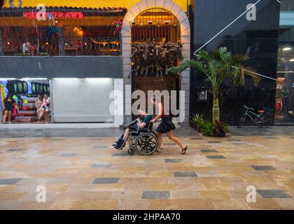 Eine lächelnde behinderte Person im Rollstuhl, die Hilfe von einem Freund erhält und durch einen tropischen Regen in einer Straße von Playa del Carmen, Mexiko, gedrückt wird Stockfoto