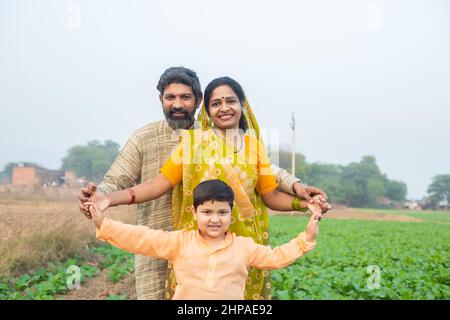 Portrait glückliche, traditionelle indische Familie auf dem landwirtschaftlichen Feld, lächelnder junger Bauer, der mit seinem Sohn und seiner Frau steht. Stockfoto