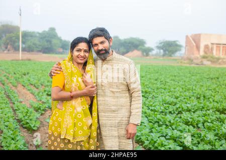 Porträt eines glücklichen indischen Landwirtspaares, das auf dem landwirtschaftlichen Feld steht, lächelnder traditioneller Mann in Kurta und Frau in Sari, die c Stockfoto