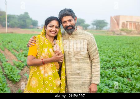 Porträt eines glücklichen indischen Landwirtspaares, das auf dem landwirtschaftlichen Feld steht, lächelnder traditioneller Mann in Kurta und Frau in Sari, die c Stockfoto