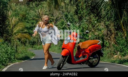 Frau auf roten Roller in weißen Kleidern fahren auf Waldweg. Tanzreise. Ein Mädchen kaukasischen Touristen in Sonnenbrillen tanzen, entspannen, ruhen in der Nähe Motorrad. Asien Thailand Reiten Tourismus. Motorradmiete. Stockfoto