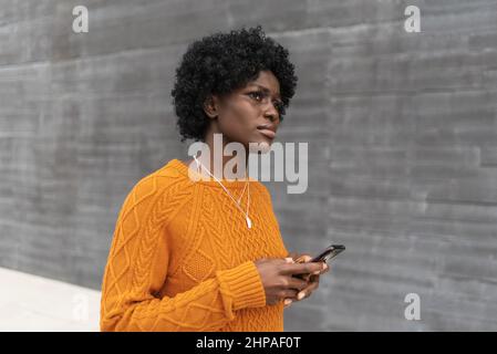 Junge Afro-Frau, die ein Mobiltelefon in der Hand hält, während sie auf der Straße läuft. Stockfoto