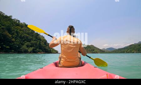 Junger Mann mit Sonnenbrille und Hut reiht sich mit einem rosa Plastikkanu entlang des Meeres gegen grüne, hügelige Inseln mit wildem Dschungel. Reisen in tropische Länder. Starker Kerl segelt auf Kajak im Ozean, Rückansicht. Stockfoto