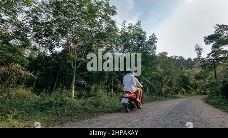 Liebe Paar auf roten Motorrad in weißen Kleidern auf Wald Road Trail Reise zu gehen. Zwei kaukasische Touristen Mann fahren auf Roller. Motorradmiete, Schutzhelm, Sonnenbrille. Asien Thailand Reiten Tourismus. Stockfoto