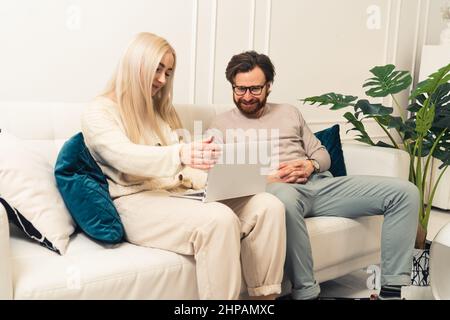 Junge kaukasische Frau mit blonden Haaren sitzt zu Hause auf der Couch und mit Laptop. In der Nähe sitzt ihr bärtiger Mann und schaut auch auf den Bildschirm. Hochwertige Fotos Stockfoto