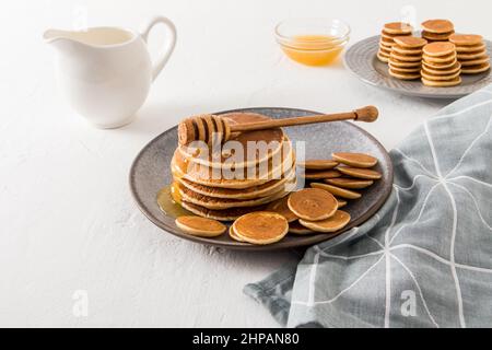 Hausgemachte Pfannkuchen und Mini-Pfannkuchen auf einem grauen Teller werden mit Honig serviert. Rahm oder Milch, eine Schüssel mit Honig, um den Geschmack zu verbessern Stockfoto