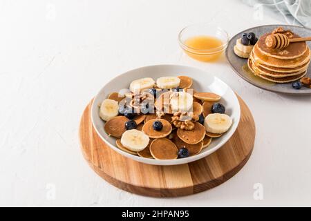 Hausgemachtes Frühstück. Eine große weiße Schüssel mit Mini-Pfannkuchen mit Bananenscheiben und Heidelbeeren auf einem Holzbrett und einem weißen Tisch Stockfoto