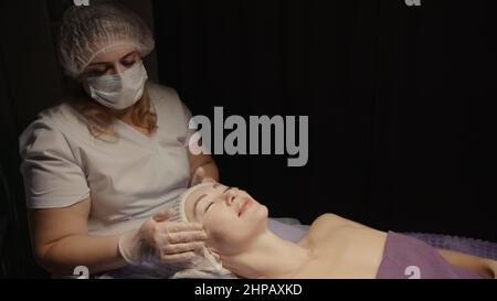 Junge hypnotische Gesichtsfrau erhält Anti-Aging-Gesichtsmassage im Spa-Salon Relax. Wellness Körper Hautpflege Gesicht Schönheitsbehandlung, Verjüngungskur bekommen Kopf Gesichtsmassage. Schwarzer Hintergrund Stockfoto