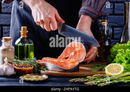 Der Küchenchef schneidet in einer Restaurantküche ein Stück rohen Lachsfischs in Scheiben. Salat, Rosmarin, Spargel, Zitrone, Flaschen und Gewürze auf einem mit Blac Stockfoto