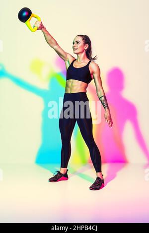 Starke Frau schwingt eine Kugelhantel. Foto der sportlichen Frau in schwarzer Sportkleidung auf weißem Hintergrund mit Effekt von rgb-Farben Schatten. Stärke und Moti Stockfoto