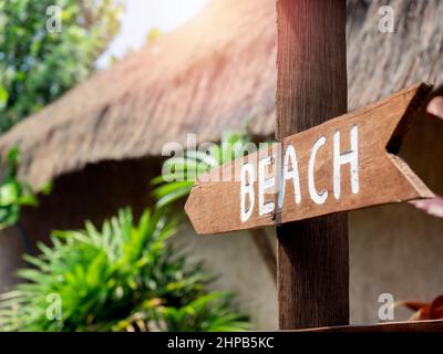 Schild mit Strandweg. Weißer Text, Strand auf der Holzbeschilderung oder Wegweiser mit Pfeilform auf dem Holzpfahl im Hotelresort. Holzzeiger zum Strand. Stockfoto